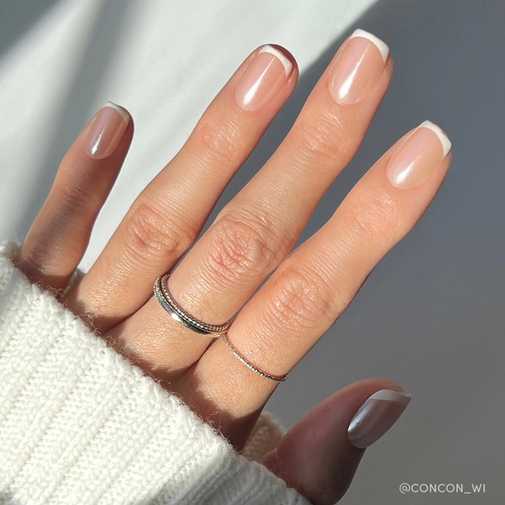 A hand with neat French tips on short squoval nails, adorned with three thin silver rings and a cozy white knit sweater—perfectly showcasing the elegant La Perle set by glamnetic.